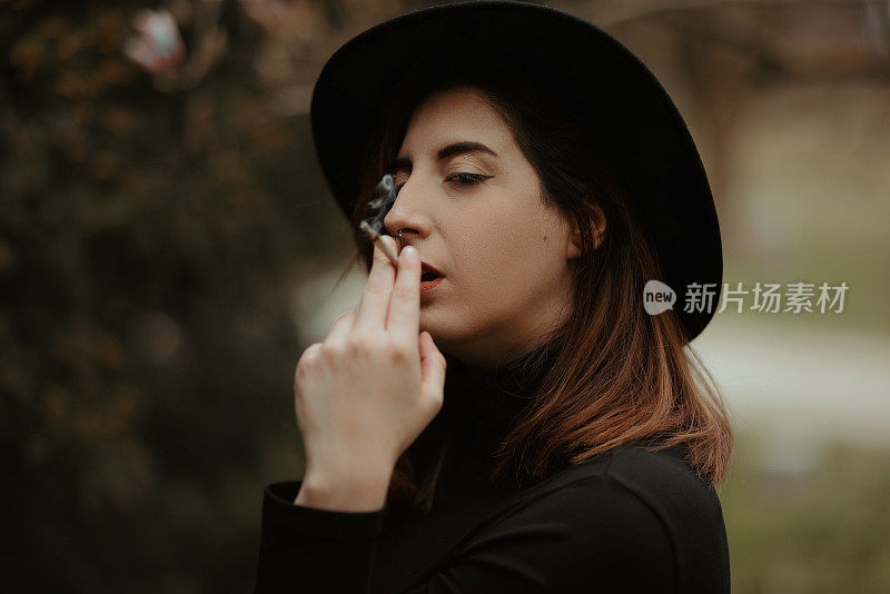 Young woman portrait wearing black hat and sweater and holding cigarette while smoking